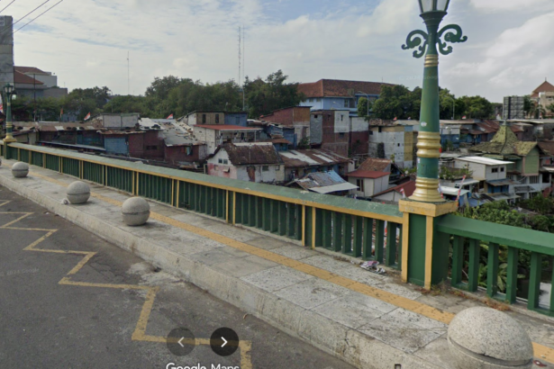 View of a city bridge with green and yellow railing, round bollards, and a river below, with densely built houses nearby.