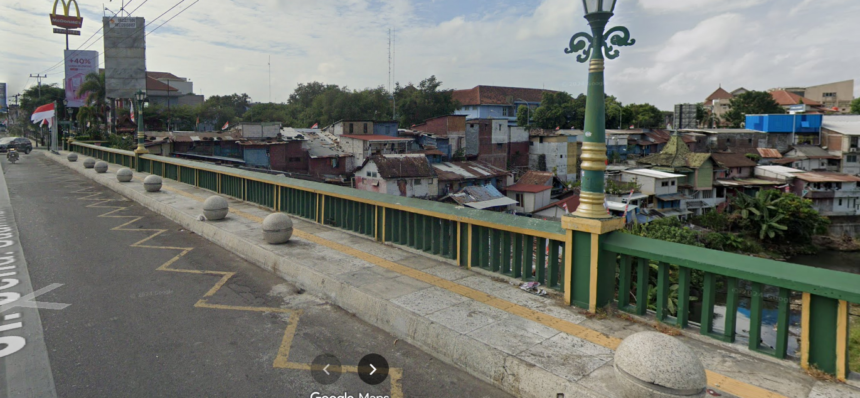 View of a city bridge with green and yellow railing, round bollards, and a river below, with densely built houses nearby.