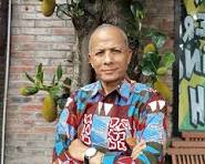 saxzaZ - Mabur.co Bald man with arms crossed in a colorful patterned shirt, standing outdoors near a brick wall and a pear tree.