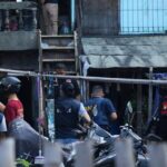 Group of police and security personnel gathered in a narrow alley outside a building, some wearing vests and helmets parked motorcycles nearby, scene of a coordinated operation.