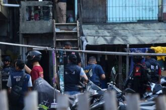 Group of police and security personnel gathered in a narrow alley outside a building, some wearing vests and helmets parked motorcycles nearby, scene of a coordinated operation.