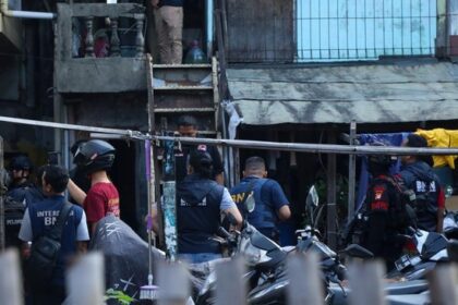 Group of police and security personnel gathered in a narrow alley outside a building, some wearing vests and helmets parked motorcycles nearby, scene of a coordinated operation.