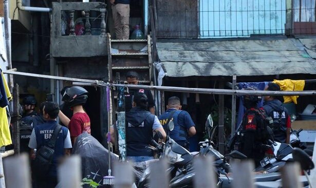 Group of police and security personnel gathered in a narrow alley outside a building, some wearing vests and helmets parked motorcycles nearby, scene of a coordinated operation.