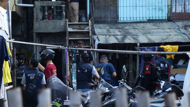 Group of police and security personnel gathered in a narrow alley outside a building, some wearing vests and helmets parked motorcycles nearby, scene of a coordinated operation.