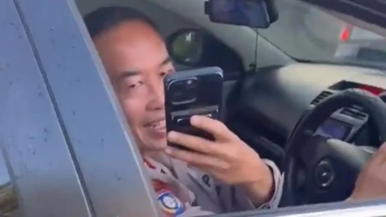 Officer in uniform taking a selfie with a cellphone inside a patrol car, reflected in the window panel.