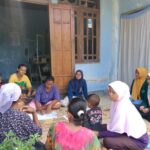 Group of women and children sit in a circle on a mat outside a blue house, likely a community gathering or lesson.