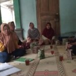 Group of women wearing hijabs sit cross-legged on a patterned mat in a circle, sharing tea and snacks in a doorway-adjacent room.