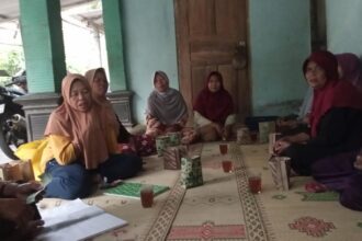 Group of women wearing hijabs sit cross-legged on a patterned mat in a circle, sharing tea and snacks in a doorway-adjacent room.
