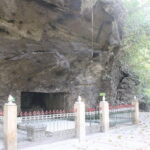 Cave entrance in a rocky cliff, surrounded by a beige fence and iron railing along a shaded walkway in a park.
