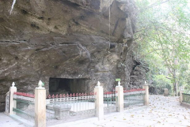 Cave entrance in a rocky cliff, surrounded by a beige fence and iron railing along a shaded walkway in a park.