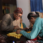 Two men in traditional clothing lean over a table, stitching yellow fabric as spectators watch.