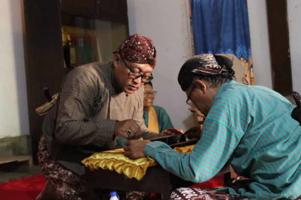 Two men in traditional clothing lean over a table, stitching yellow fabric as spectators watch.