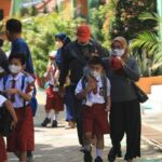 Group of schoolchildren in red uniforms with backpacks walking outdoors with supervising adults, all wearing masks in a sunny campus courtyard