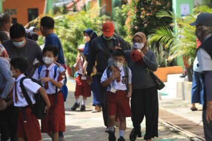 Group of schoolchildren in red uniforms with backpacks walking outdoors with supervising adults, all wearing masks in a sunny campus courtyard