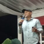 Man in a light shirt and cap speaks into a handheld microphone on a stage, holding a phone in his other hand.