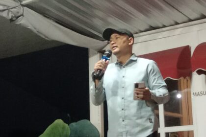 Man in a light shirt and cap speaks into a handheld microphone on a stage, holding a phone in his other hand.