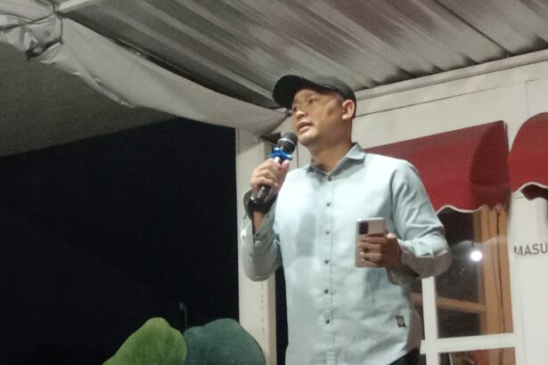 Man in a light shirt and cap speaks into a handheld microphone on a stage, holding a phone in his other hand.