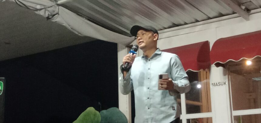 selasa sastra juli 2026 2 - Mabur.co Man in a light shirt and cap speaks into a handheld microphone on a stage, holding a phone in his other hand.