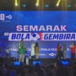 Stage event with a large blue banner reading SEMARAK BOLA GEMBIRA; hosts and participants stand on stage with drums and audience in foreground.