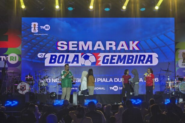 Stage event with a large blue banner reading SEMARAK BOLA GEMBIRA; hosts and participants stand on stage with drums and audience in foreground.