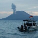 Small motorboat with people near a volcanic island emitting a smoke plume; an Indonesian flag flies at the stern.