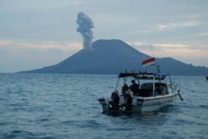 Small motorboat with people near a volcanic island emitting a smoke plume; an Indonesian flag flies at the stern.