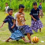 Group of barefoot young boys playing soccer on a grassy field, wearing colorful wraparound skirts and chasing a yellow-and-pink ball.