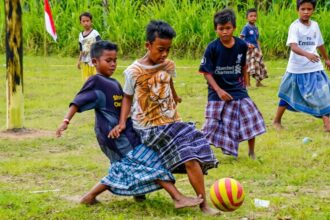 Group of barefoot young boys playing soccer on a grassy field, wearing colorful wraparound skirts and chasing a yellow-and-pink ball.