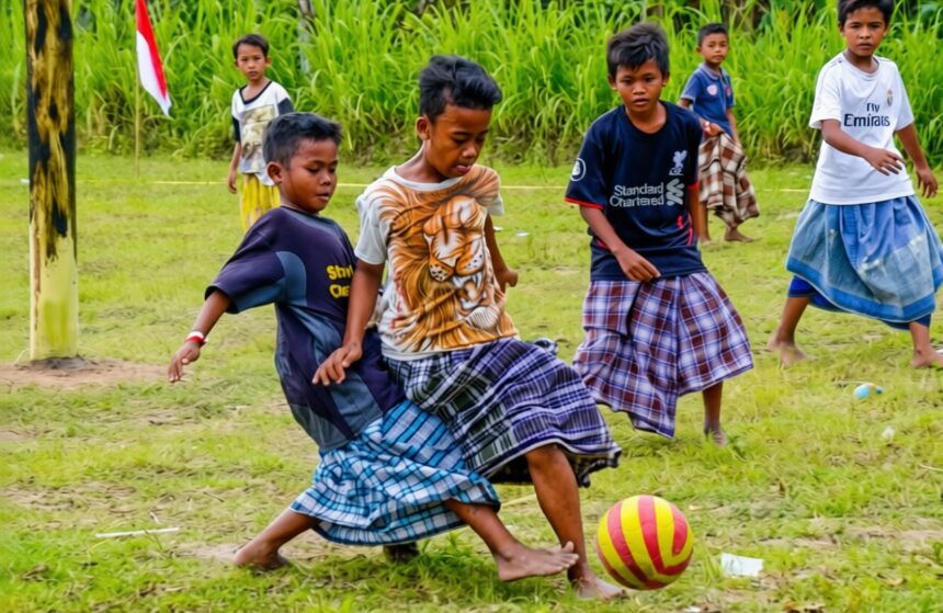 sepakbola sarung by antara foto FIXX - Mabur.co Group of barefoot young boys playing soccer on a grassy field, wearing colorful wraparound skirts and chasing a yellow-and-pink ball.
