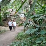 Two men in white shirts walk along a dirt path through dense tropical jungle with large green leaves surrounding them.