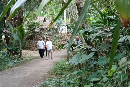 Two men in white shirts walk along a dirt path through dense tropical jungle with large green leaves surrounding them.