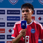 Soccer player in a red jersey being interviewed, holding a blue Hyundai microphone, with sponsor backdrop behind him.