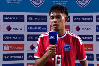 Soccer player in a red jersey being interviewed, holding a blue Hyundai microphone, with sponsor backdrop behind him.