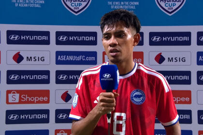 Soccer player in a red jersey being interviewed, holding a blue Hyundai microphone, with sponsor backdrop behind him.