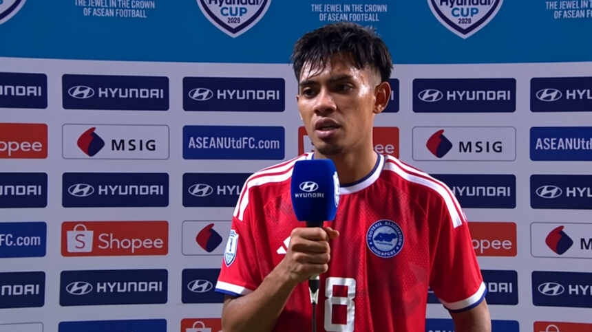 Soccer player in a red jersey being interviewed, holding a blue Hyundai microphone, with sponsor backdrop behind him.