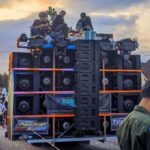 A large, multi‑stacked speaker system mounted on a flatbed truck with people on top during a street parade; Indonesian flags line the street in the background.