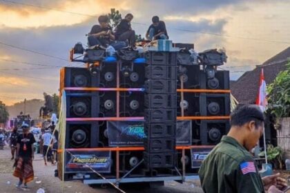 A large, multi‑stacked speaker system mounted on a flatbed truck with people on top during a street parade; Indonesian flags line the street in the background.