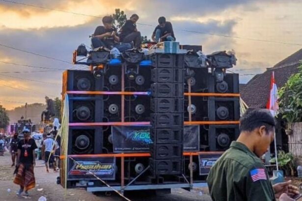 A large, multi‑stacked speaker system mounted on a flatbed truck with people on top during a street parade; Indonesian flags line the street in the background.