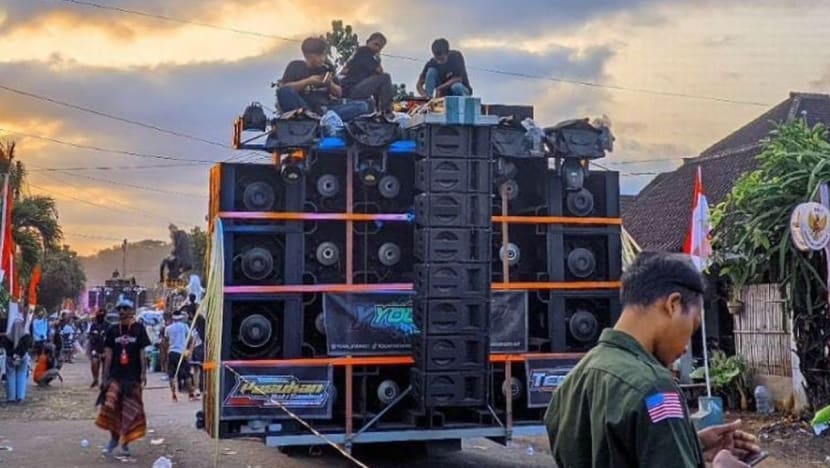 A large, multi‑stacked speaker system mounted on a flatbed truck with people on top during a street parade; Indonesian flags line the street in the background.