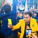 Man in a bright yellow blazer with glasses speaks into a microphone while raising a yellow card amid a large crowd at a rally.