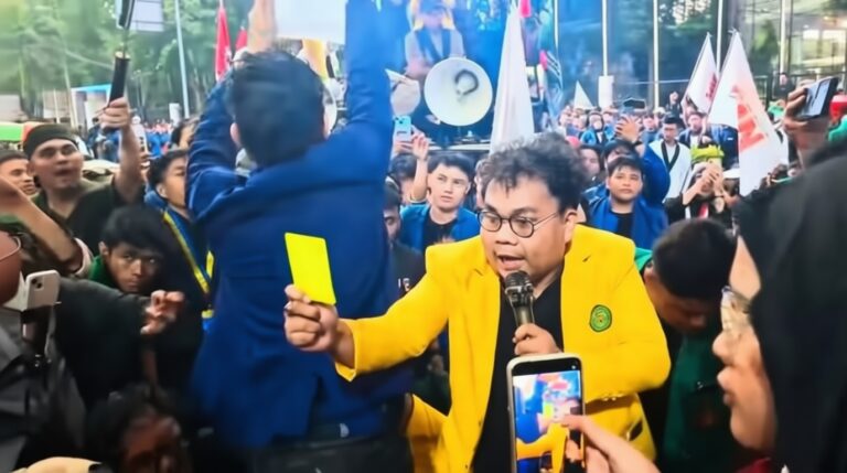 Man in a bright yellow blazer with glasses speaks into a microphone while raising a yellow card amid a large crowd at a rally.
