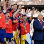 Group of athletes in red jerseys celebrate with medals as two suited men clap nearby in a stadium crowd.