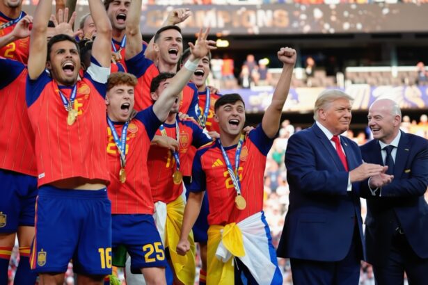 Group of athletes in red jerseys celebrate with medals as two suited men clap nearby in a stadium crowd.