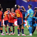 Spain national team players in red and blue jerseys hydrate with water bottles during a match break on the field.