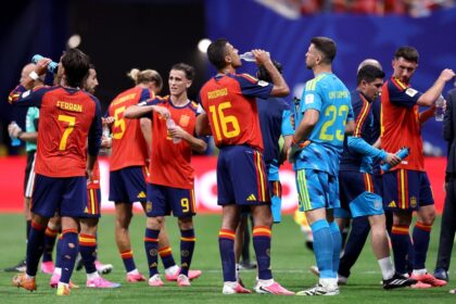 Spain national team players in red and blue jerseys hydrate with water bottles during a match break on the field.