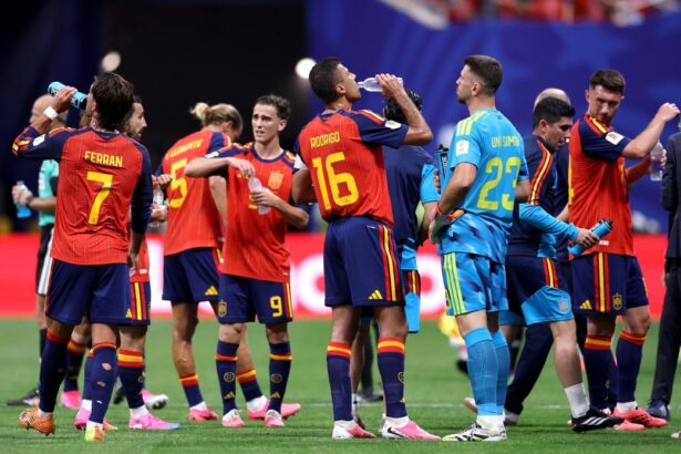 Spain national team players in red and blue jerseys hydrate with water bottles during a match break on the field.