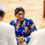 Person wearing a bright blue floral traditional outfit and black head wrap sits with hands folded, listening in a warmly lit room among blurred others in the foreground.