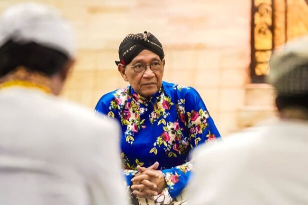 Person wearing a bright blue floral traditional outfit and black head wrap sits with hands folded, listening in a warmly lit room among blurred others in the foreground.