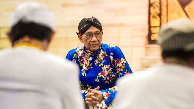Person wearing a bright blue floral traditional outfit and black head wrap sits with hands folded, listening in a warmly lit room among blurred others in the foreground.