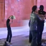 A barefoot person in a black top raises a white board toward a masked figure as three men approach near a pink-tiled wall with 'ART JOG' on the left.